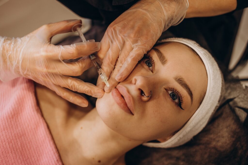 Woman lying down with eyes wide open as beauty professional wearing gloves inserts needle into her lips.