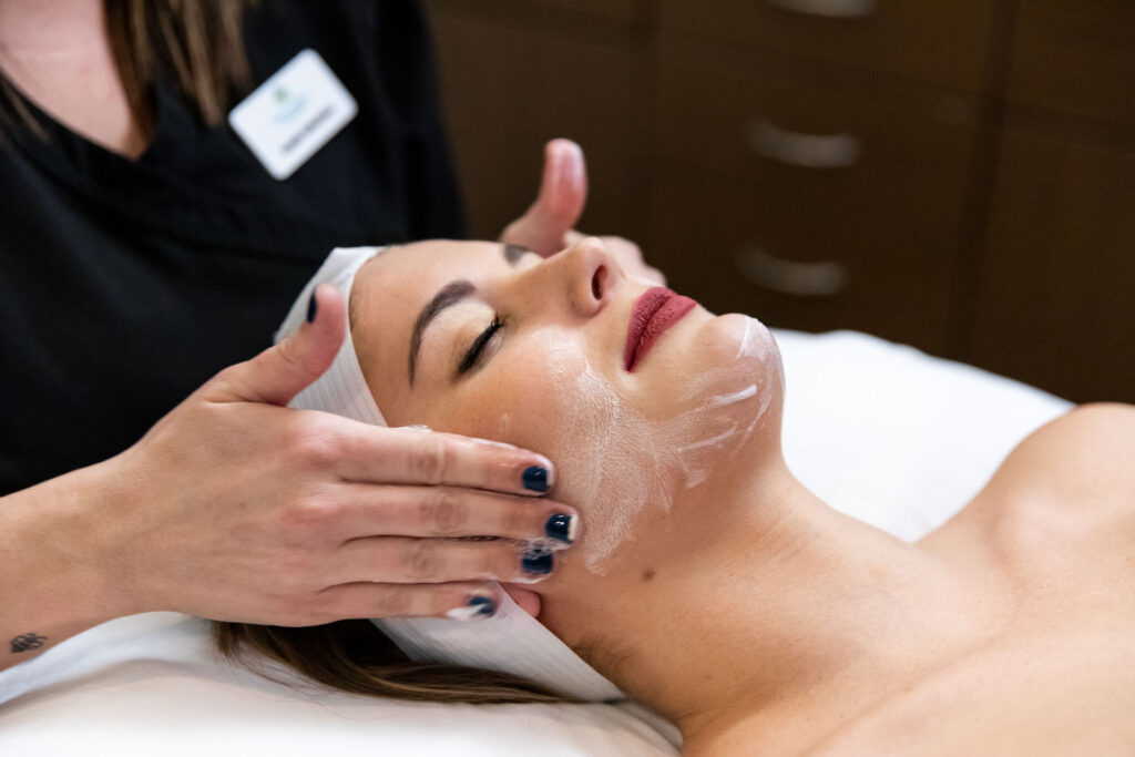 Side shot of woman lying down while professional massages facial cream on her cheeks.
