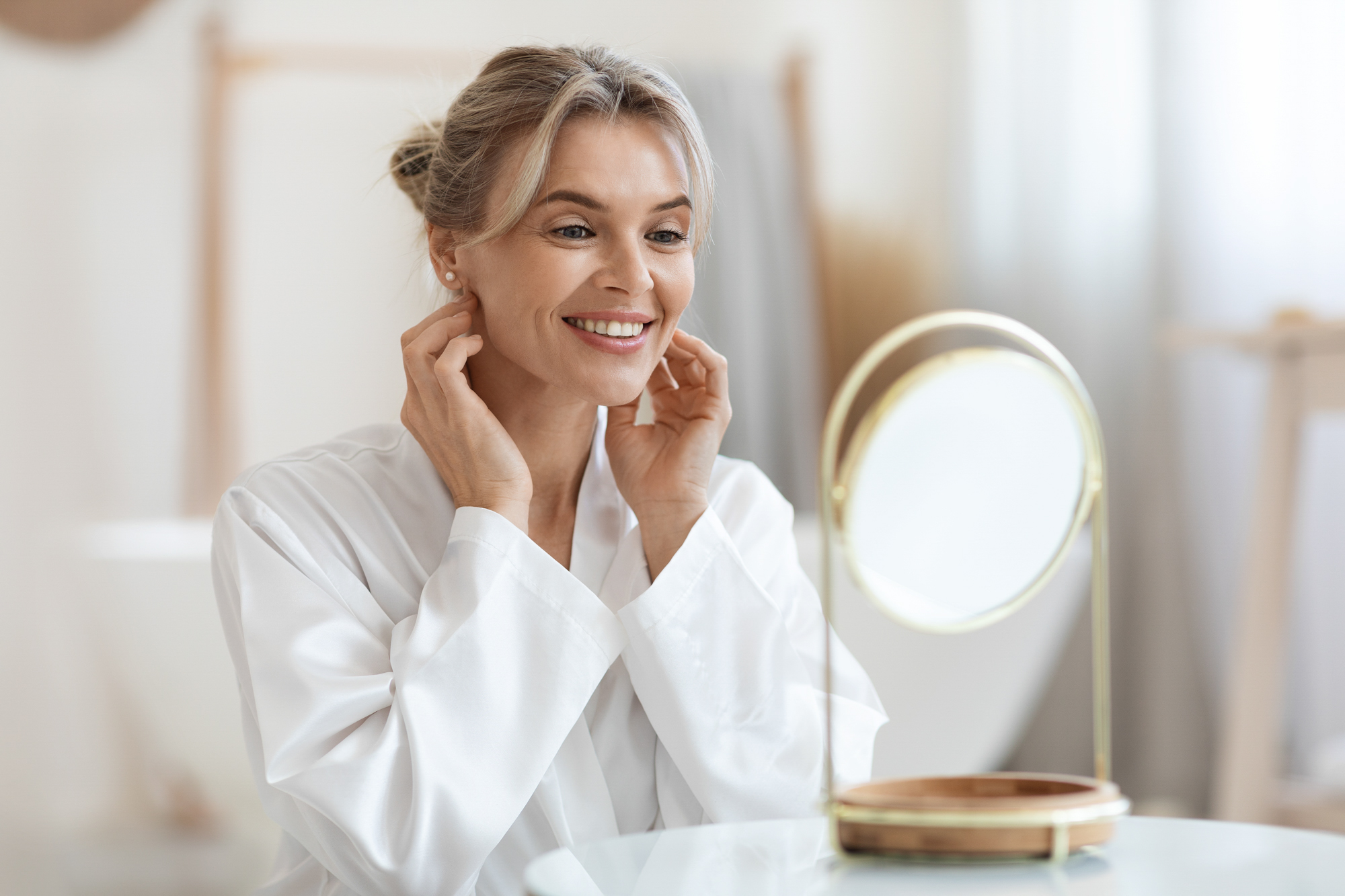 A blonde woman at home looking happily at her reflection after visiting the team at Exhibit Medical Aesthetics for injections of Botox near Madison, NJ.