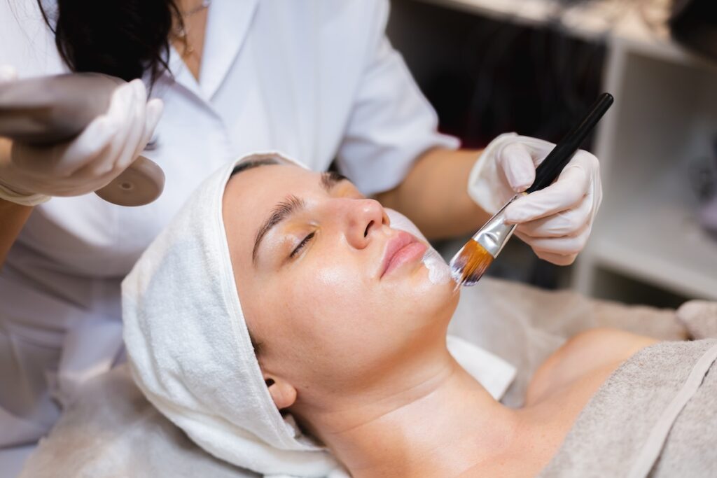 Woman lying down with eyes closed as beautician applies mask with brush.