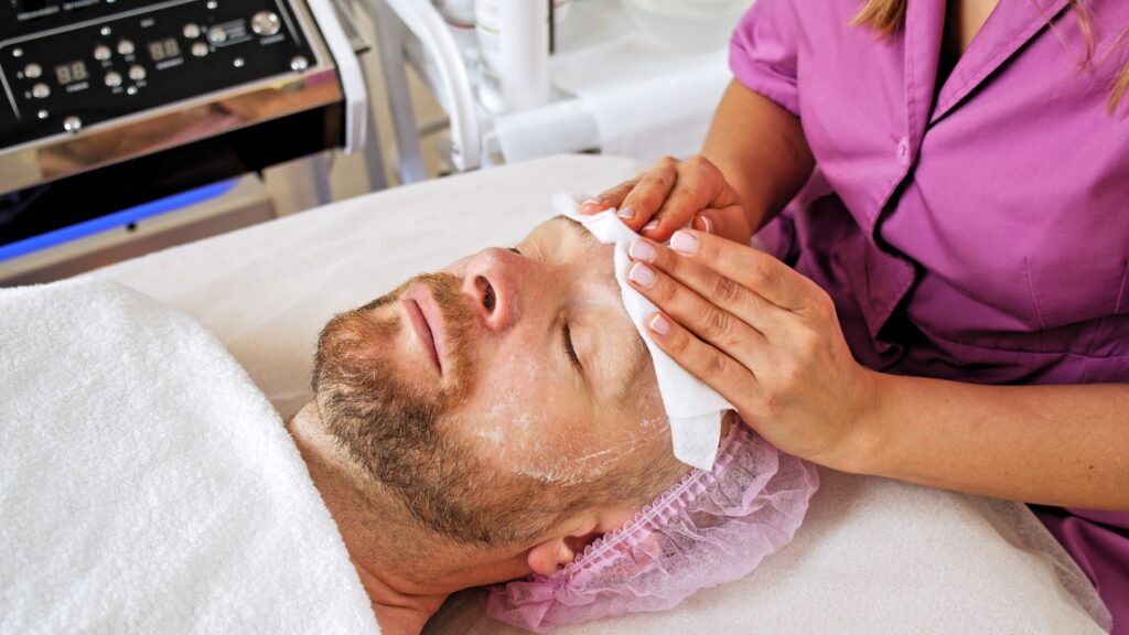 Cosmetologist holds cloth on man's face as he lies down.