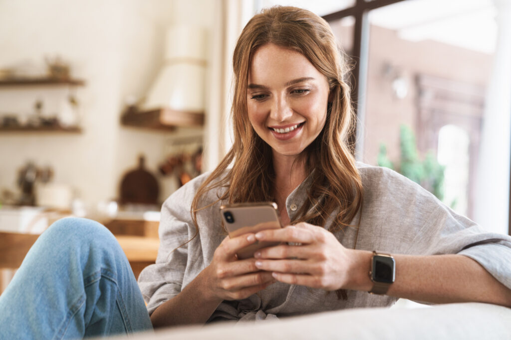 A young woman sitting on her couch at home, looking on her phone at the Exhibit Medical Aesthetics website. She is specifically looking at getting chin filler near Mountain Lakes, NJ.