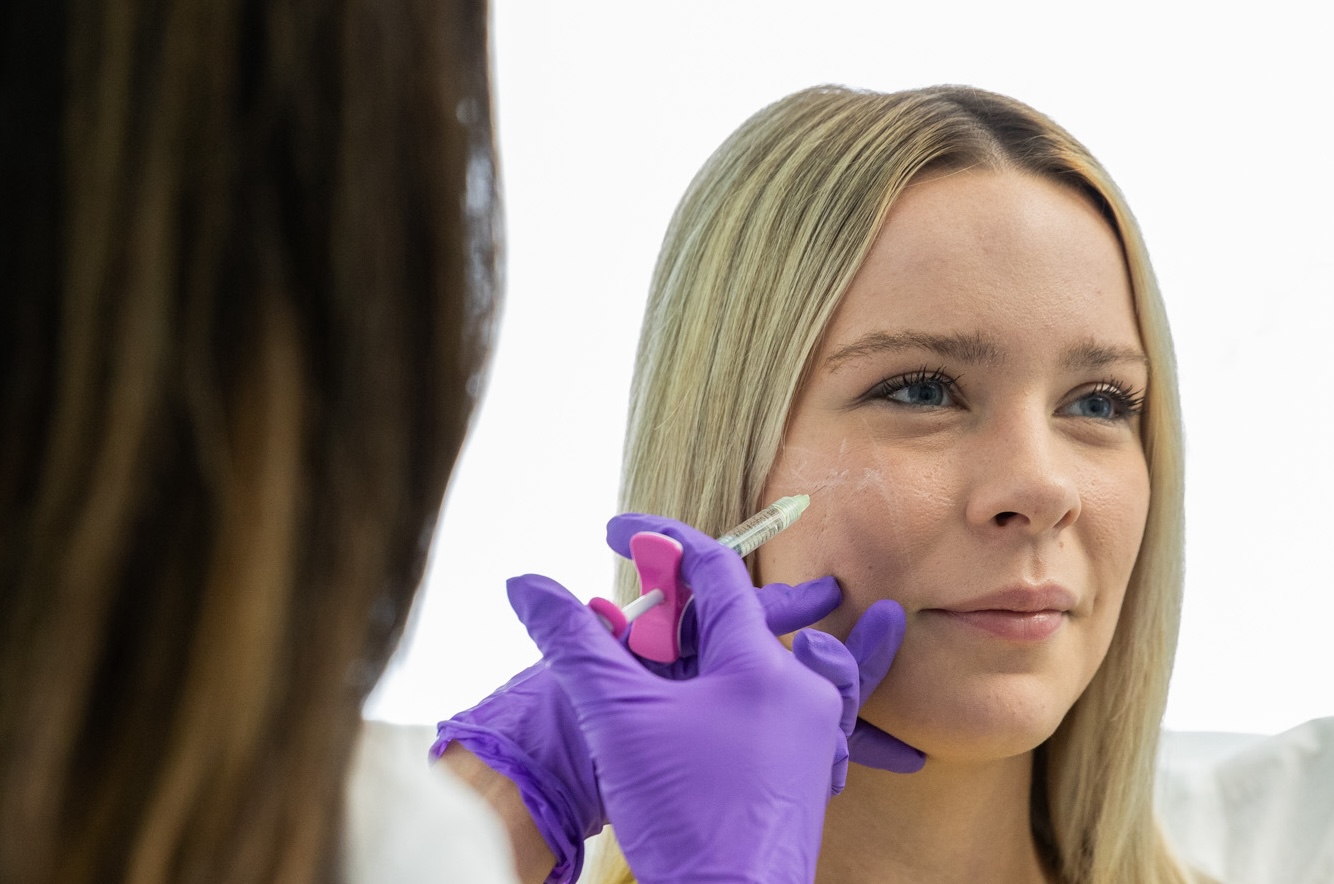 A young blonde patient sitting comfortably in one of the treatment rooms at Exhibit Medical Aesthetics while an expert injector administers her filler near Madison, NJ.
