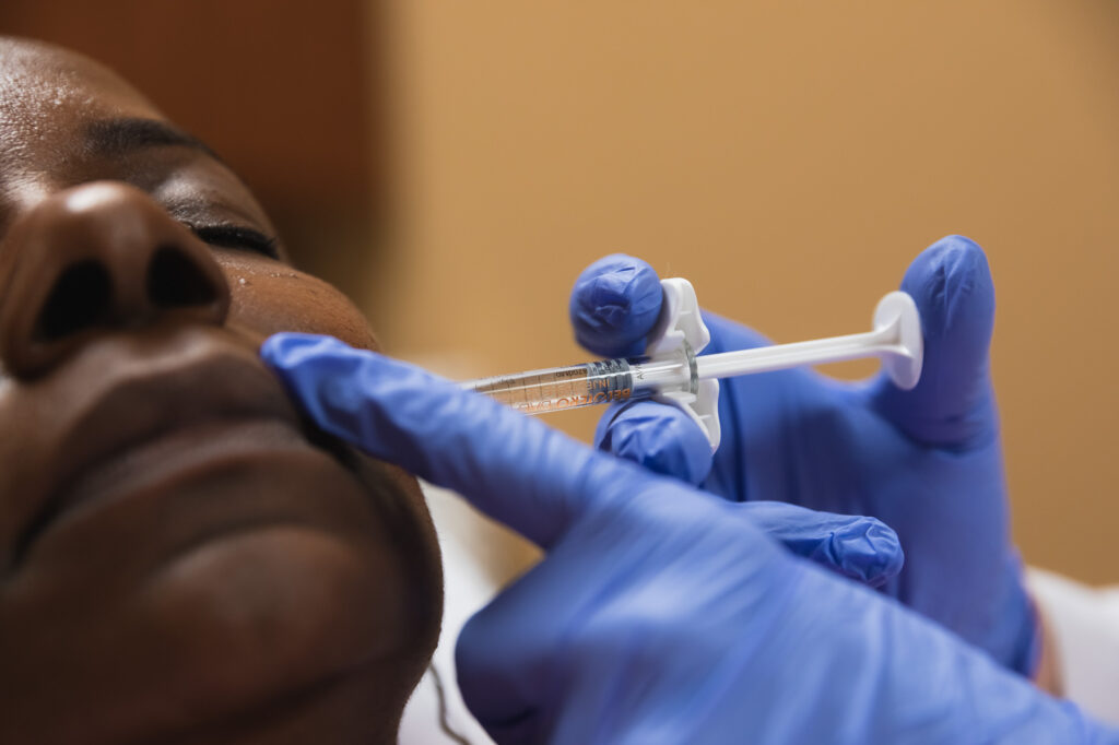 Patient closes eyes as professional wearing blue gloves inserts filler into the side of her face.