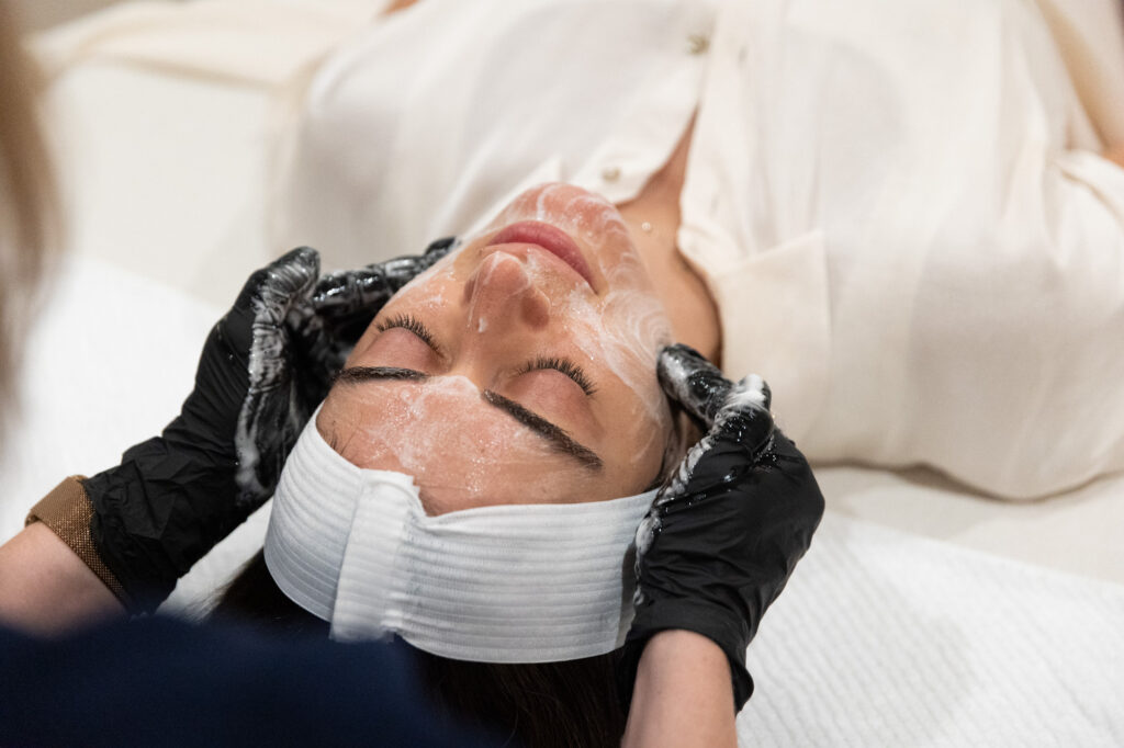 Woman with eyes closed as professional wearing gloves massages her face.