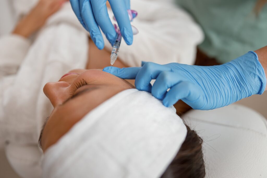 Beautician wearing blue gloves injects needle into patient's face as she's laying down.